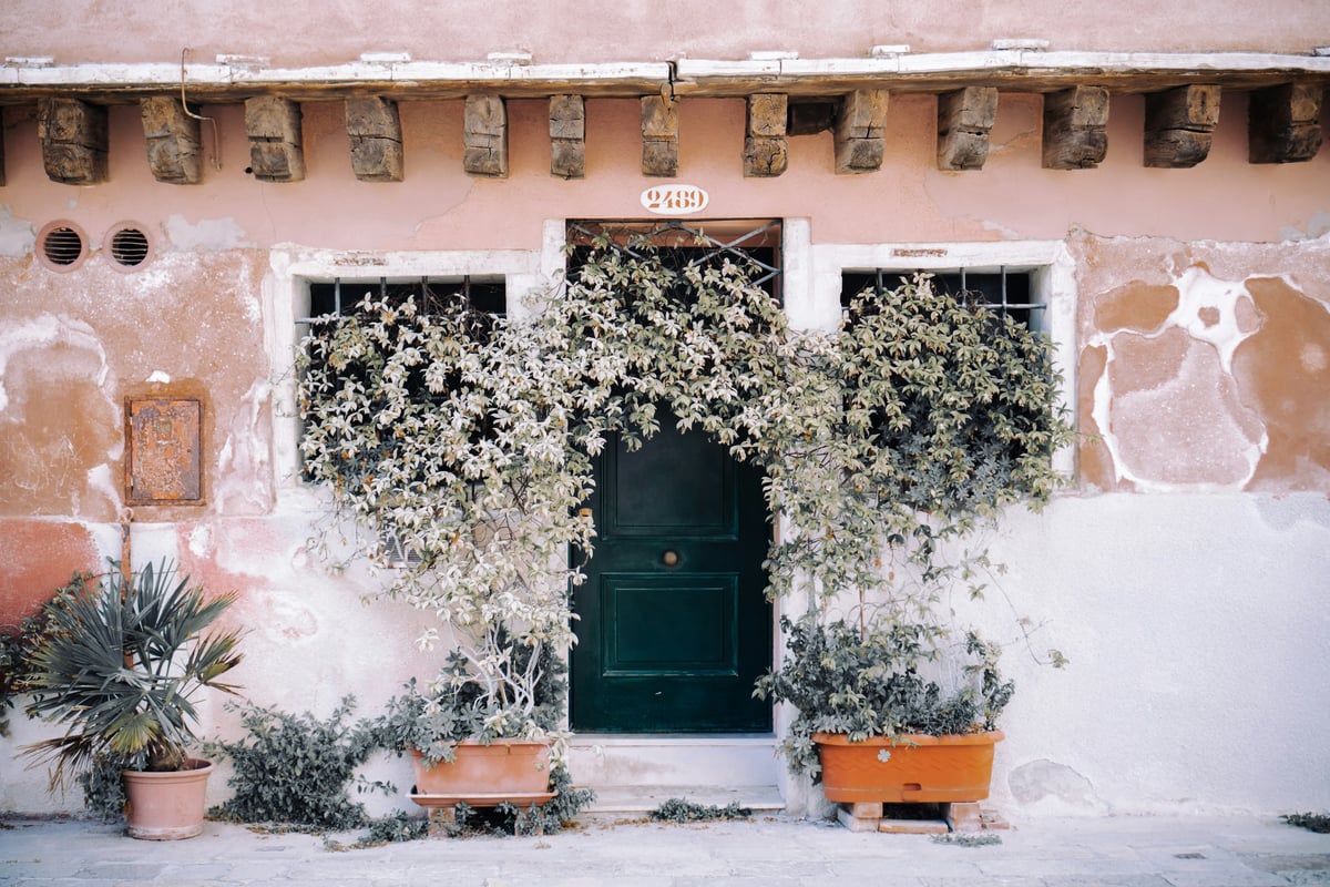 A charming Italian building entrance with a green door and plants, representing the kind of home you could find after relocating to Italy with Why Wait Italy.