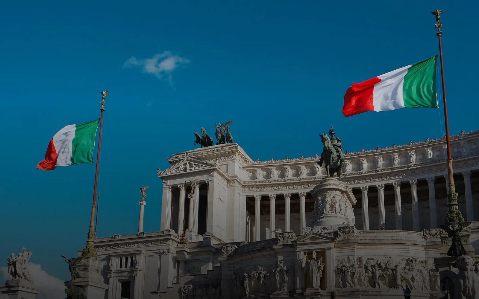 Two Italian flags fly in front of the white marble colonnade and equestrian statue of the Vittoriano in Rome under a deep blue sky.