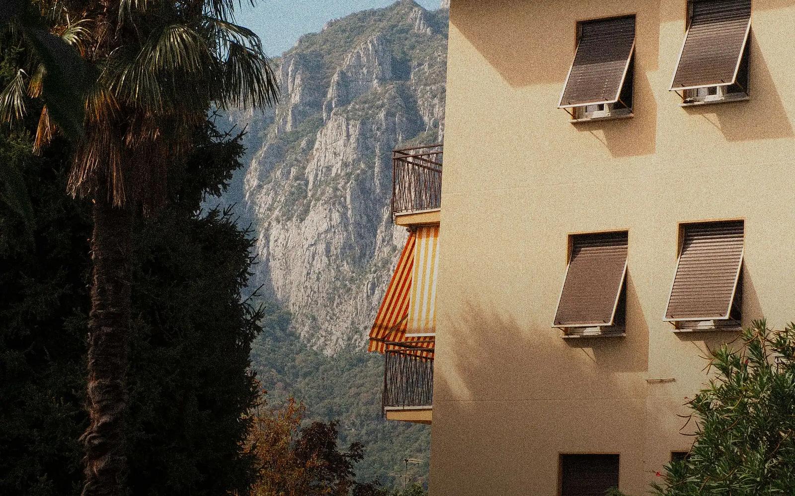 An apartment block in Lecco with open shutters and a striped balcony awning, a palm tree in front and the limestone face of the mountain behind.