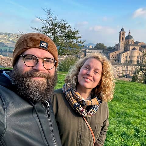 A smiling couple takes a selfie on a grassy hill overlooking a picturesque Italian town.