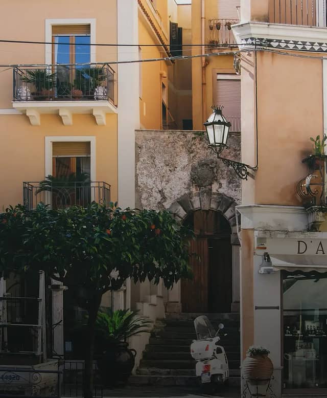A sunlit Sicilian street with orange-washed apartment buildings, balconies, an orange tree and a Vespa parked by a stone stairway.