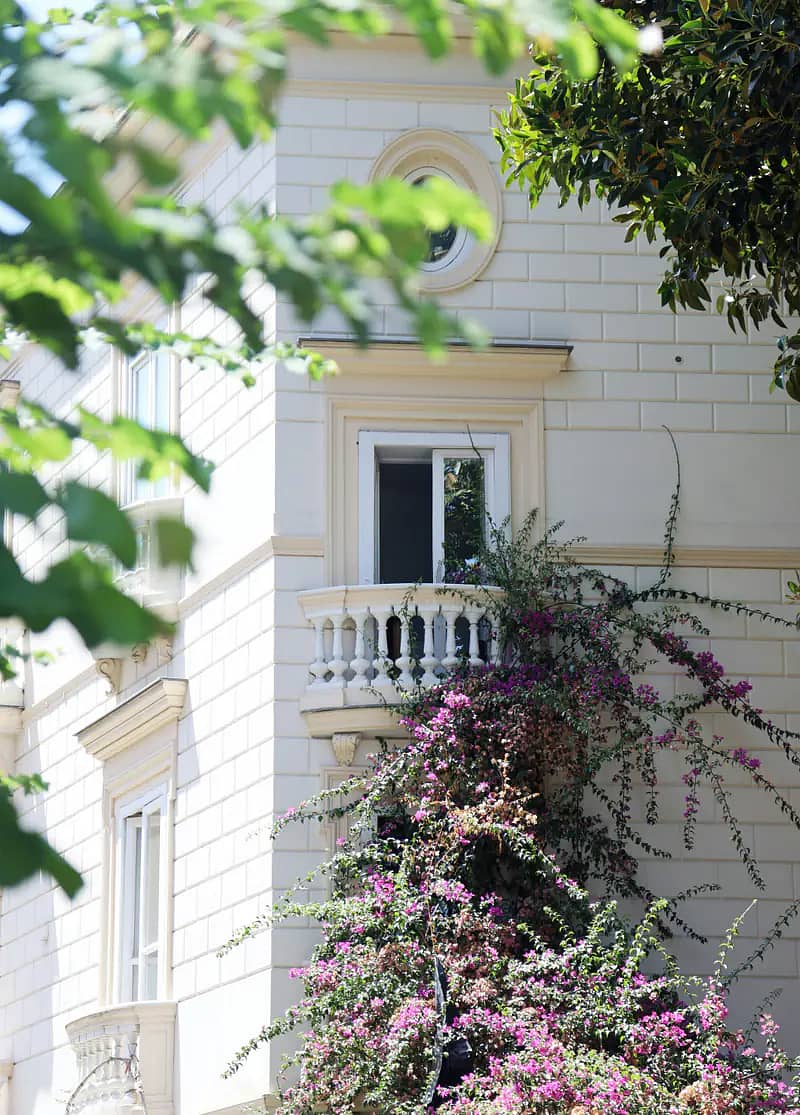 A pale stone villa with an oval window and a small balustraded balcony, pink bougainvillea climbing the wall below it, seen through leaves.