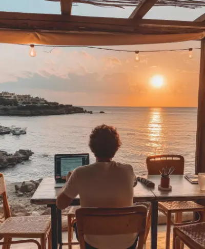 A digital nomad works on a laptop at a table overlooking a sunset ocean view.