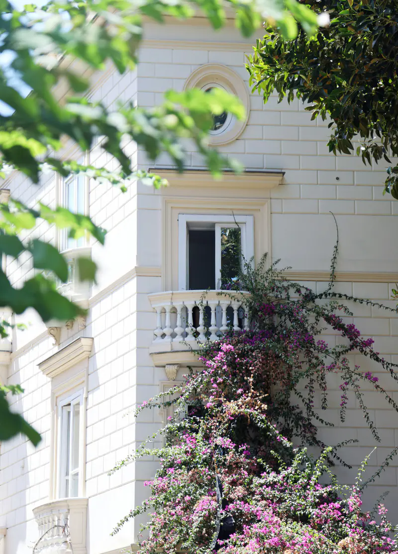 A pale stone villa with an oval window and a small balustraded balcony, pink bougainvillea climbing the wall below it, seen through leaves.