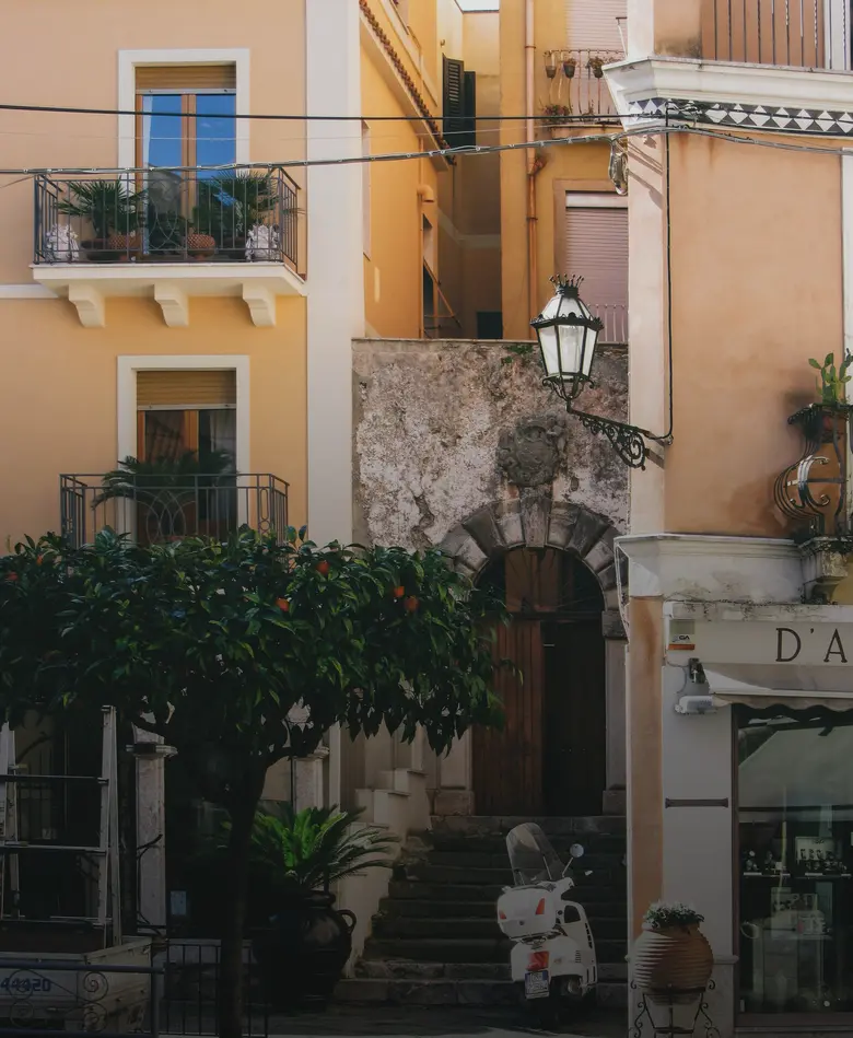 A sunlit Sicilian street with orange-washed apartment buildings, balconies, an orange tree and a Vespa parked by a stone stairway.